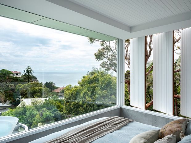 A bright coastal bedroom corner with a bed positioned beside expansive corner glazing. The room has a white-painted timber-lined ceiling, pale grey walls and window framing, vertical white privacy screens, and views over leafy residential surroundings toward the ocean. Neutral bedding and cushions create a calm, airy atmosphere, while mature trees are visible immediately outside.