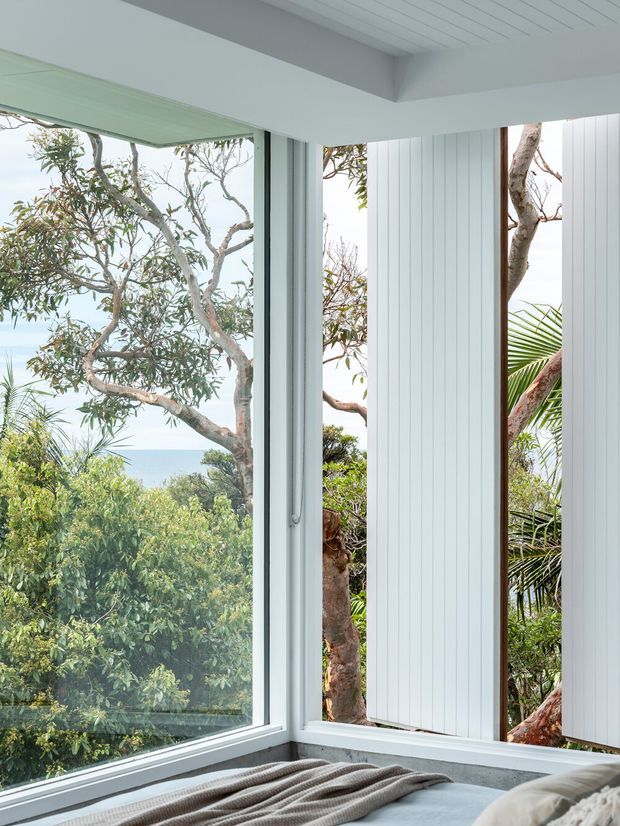 Bright coastal bedroom corner with a low bed or daybed in the foreground, framed by expansive corner glazing. White vertical wall panelling and a white slatted ceiling create a clean, airy interior, while lush trees, palms, and a glimpse of blue water are visible outside. The space has a calm, minimal coastal character.