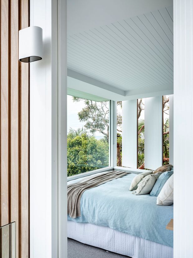 A bright coastal-style bedroom viewed through an open doorway, featuring a pale blue bedspread, layered neutral bedding, large corner windows overlooking greenery, and a white vertically panelled ceiling and walls. The doorway includes warm timber detailing and a simple white wall light. The room has abundant natural light and a strong connection to the surrounding landscape.
