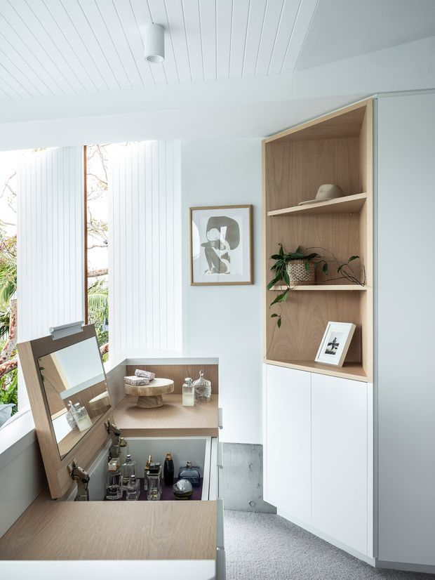 Bright, refined dressing and wardrobe area with white vertical-panel walls and ceiling, large windows, pale carpet, and a built-in white-and-light-oak display cabinet. The open shelving holds decorative objects and plants, while a mirrored vanity or dressing cabinet with integrated storage occupies the foreground. The room has a calm, minimalist contemporary feel with abundant natural light.