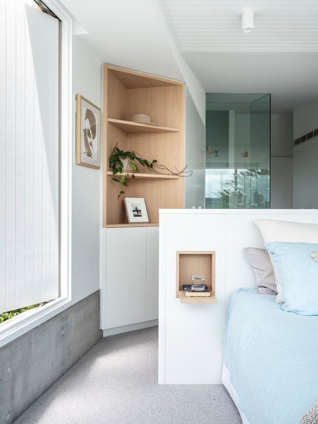 Bright, compact contemporary bedroom with a pale blue bed in the foreground, built-in white cabinetry and open light-wood shelving, a small bedside niche, framed artwork, and a large window. A glass-partitioned bathroom or shower area is visible beyond the cabinetry. The room has a clean, minimal fit-out with soft natural light and neutral finishes.