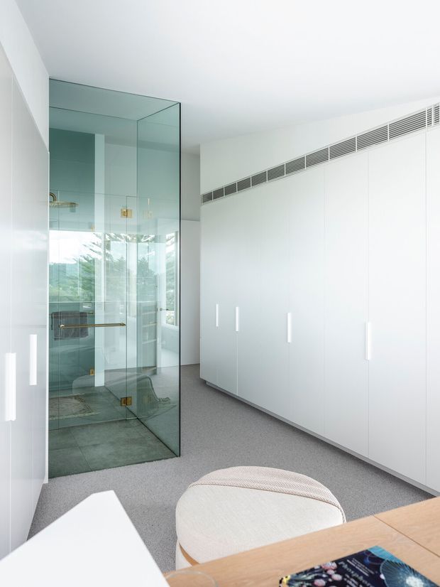 Bright minimalist dressing room or wardrobe area with extensive full-height white built-in cabinetry, slim integrated handles, and grey carpet flooring. A transparent glass-enclosed bathroom or shower zone is visible to the left, with pale green-grey tiled surfaces and brass hardware. The space has a clean contemporary fit-out, concealed linear air-conditioning vents, and abundant natural light through glazing. A rounded upholstered stool and timber desk or bench appear in the foreground.