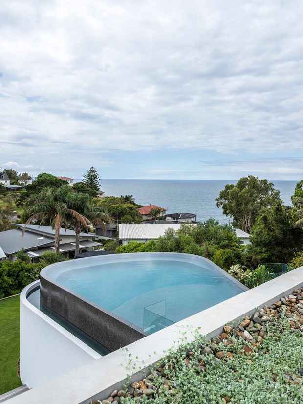 Elevated exterior view of a modern rooftop or infinity-edge swimming pool overlooking a coastal residential neighbourhood and the ocean. The pool has dark textured sides, pale concrete coping, and a clear glass safety panel, with a planted rooftop garden and landscaped greenery in the foreground. The scene is bright but overcast, with a calm ocean horizon and contemporary houses visible beyond.