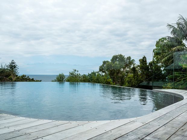 An infinity-edge swimming pool on a timber deck overlooks a coastal or ocean horizon, surrounded by dense tropical and subtropical vegetation. The pool has a curved edge and the scene is viewed from the deck under an overcast sky.