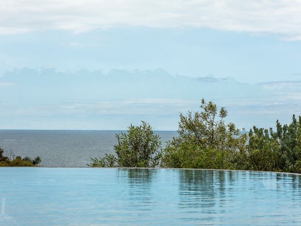 A tranquil infinity pool fills the foreground, its water visually merging with the calm ocean horizon beyond. Low coastal vegetation frames the pool edge under a softly overcast blue sky. The image emphasizes the seamless connection between the pool and seascape, with no building or interior visible.