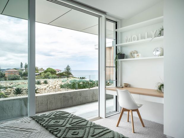 Bright, contemporary bedroom with a built-in white study desk and open shelving, viewed beside full-height glazed doors leading to a balcony or terrace. The room has a textured grey carpet, a bed with a geometric throw, white shelving and joinery, and a white moulded chair with timber legs. Outside is a planted coastal landscape and ocean view, with a concrete planter wall and glass balustrade.