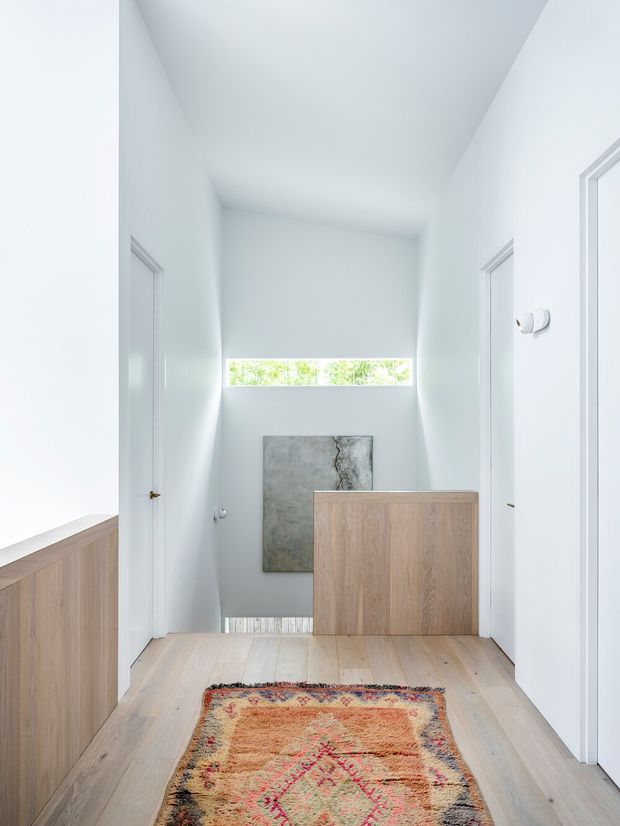 Bright minimalist residential hallway or landing with white walls, a high angled ceiling, pale timber flooring, flush white doors, and light timber built-in cabinetry. A narrow horizontal clerestory window brings in daylight, while a framed artwork and patterned runner add subtle warmth. A stair opening or lower-level transition is visible at the far end.
