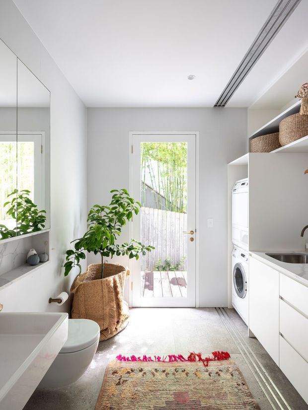 Bright, narrow contemporary laundry-bathroom utility space with a stacked front-loading washer and dryer, white built-in cabinetry, open shelving with woven baskets, a deep sink, toilet and partial bathtub or basin. A full-height mirrored wall expands the room visually, while a glazed exterior door opens to a small timber deck and fenced garden. The space uses pale grey walls, white joinery, light flooring and natural woven accents, with linear recessed ceiling lighting.