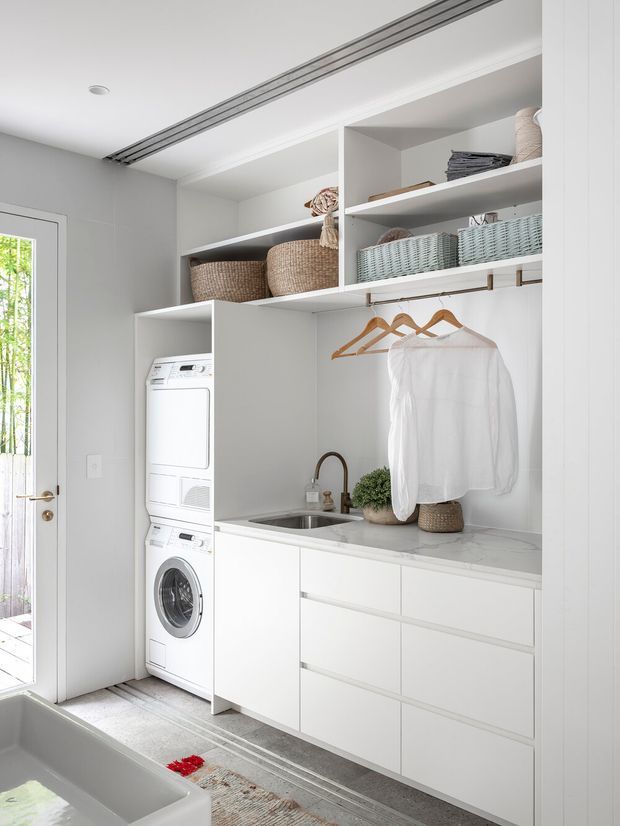 Bright, compact laundry room with white built-in cabinetry, stacked washer and dryer, open shelving with woven baskets, hanging rail, utility sink, and pale stone-look benchtop. A glazed exterior door provides natural light, while a bathtub edge is visible in the foreground, suggesting the laundry is adjacent to a bathroom or utility area.