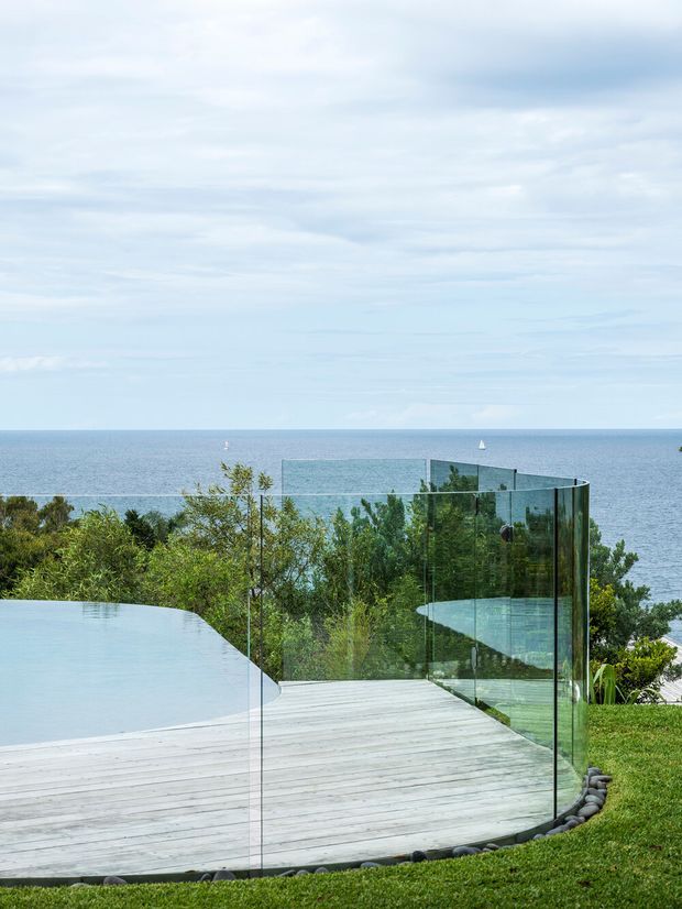 Exterior coastal view of a contemporary property with a curved glass balustrade enclosing a pale timber deck and infinity-edge pool. Dense landscaped vegetation surrounds the deck, with an expansive ocean horizon and sailboats beyond. The image is bright, open, and strongly connected to the seaside setting.