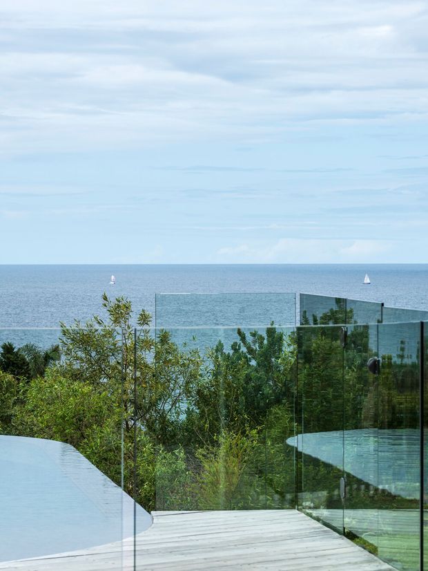 A coastal outdoor view featuring a pale timber deck beside a curved pale-blue pool, bordered by transparent glass balustrades. Dense coastal planting sits between the pool and the ocean, with open sea and sailboats visible beyond under a lightly clouded sky.