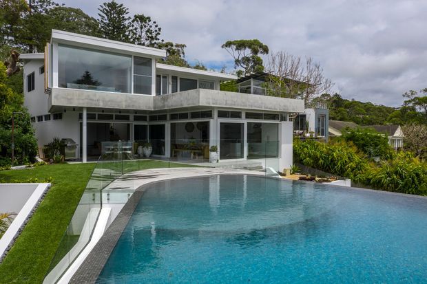A contemporary multi-level residential house viewed from the swimming pool. The home features extensive floor-to-ceiling glazing, white and grey concrete forms, projecting upper-level volumes, and a broad balcony overlooking the pool. Glass pool fencing, manicured lawn, and dense landscaped planting surround the outdoor pool area.