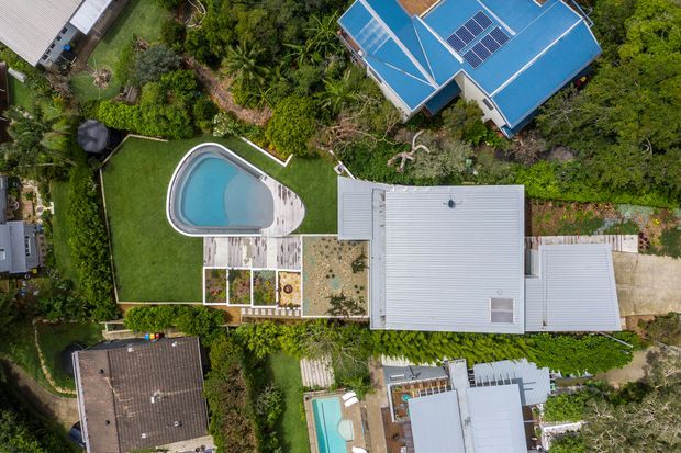 Aerial top-down view of a leafy residential property with several buildings, light-coloured metal roofs, a curved swimming pool, paved outdoor areas, lawns, dense tropical landscaping, and rooftop solar panels visible on the upper building.