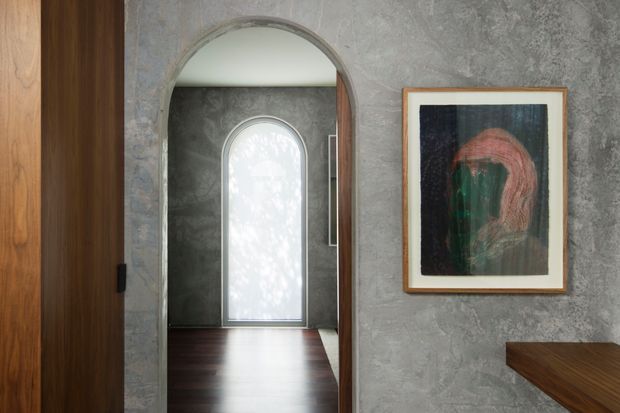 A refined interior hallway or entry passage with a tall arched opening and arched frosted window beyond. The walls have a textured grey finish, paired with dark timber joinery and flooring. A framed abstract artwork hangs on the right wall, while a dark timber surface enters the lower-right corner. The composition is quiet, warm, and architectural, with strong curved forms and natural timber accents.