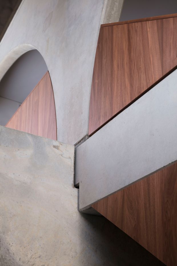 Close-up architectural view of a sculptural stairway with angular timber-clad elements intersecting pale textured plaster or concrete walls. A rounded arch opening is visible to the left, contrasting with the sharply geometric stair and balustrade forms. The composition is minimalist and material-focused, with warm walnut-toned timber against cool light-grey surfaces.