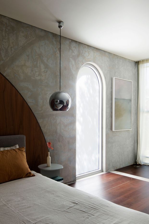 A refined bedroom interior with a bed in the foreground, a curved dark timber feature wall and integrated bedside area, mottled grey decorative plaster walls, a tall arched glazed door or window, and dark timber flooring with a light inlay strip. A chrome spherical pendant hangs beside the bed, complemented by sheer curtains and minimalist abstract artwork. The room has a warm, sculptural, contemporary character.