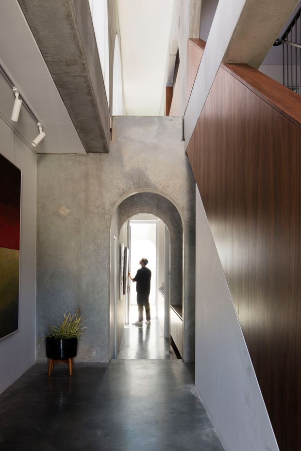 A dramatic interior hallway with a polished dark concrete floor, raw concrete walls and ceiling elements, and a rounded concrete arch leading toward a bright doorway. A large dark timber built-in stair or joinery volume lines the right side, with black metal stair railings above. Track lighting, a potted plant, and framed artwork add restrained accents. The space has strong vertical lines, contrasting materials, and a high, light-filled architectural composition.