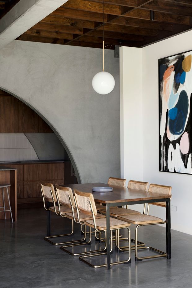 A sophisticated dining area with a long dark timber dining table and matching cantilevered woven-seat chairs. The room features a polished grey concrete floor, a curved exposed concrete architectural wall framing a compact kitchen or bar area, dark timber ceiling beams and panelling, a white painted wall, and a bold abstract artwork. A spherical pendant light hangs centrally over the table, creating a refined modern-industrial atmosphere.