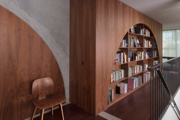 Interior view of a warm, wood-lined landing or hallway featuring a large curved built-in bookshelf filled with books, a sculptural timber chair, a textured concrete arch detail, dark timber flooring, and a black metal stair balustrade. Soft daylight enters from glazing at the far end.