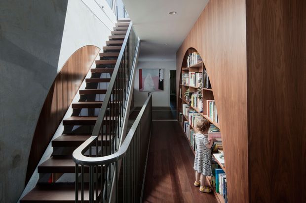 A contemporary interior hallway and stairway with a dramatic open-riser staircase, dark timber treads, slim dark metal balustrade, and a curved white handrail. Full-height timber wall panelling and integrated arched bookcases line the corridor, with dark timber flooring and artwork at the far end. A child is present near the shelving, but the focus is the architectural circulation space and built-in joinery.