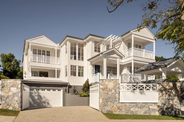 Exterior view of a large multi-storey white weatherboard residence with prominent gabled roofs, multiple covered balconies, white decorative railings and verandahs, stone boundary walls, and an integrated garage. The home has a refined coastal/traditional appearance and sits on a landscaped suburban street frontage.