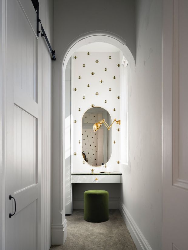 A narrow, bright dressing and vanity area viewed through an arched opening. The space features white panelled walls and doors, a floating vanity with a pale stone benchtop, an oval mirror, ornate gold wall-mounted lighting, patterned wallpaper with small gold motifs, and a green upholstered stool on a grey carpet.