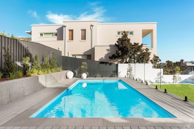 A contemporary multi-level home with clean white geometric volumes, narrow vertical windows, and a large open balcony or void. The foreground features a bright rectangular swimming pool surrounded by grey decking, concrete retaining walls, dark vertical fencing, landscaped planting, and a manicured lawn under a clear blue sky.