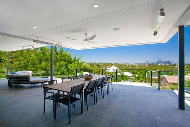 A spacious covered outdoor terrace with a long dining table and chairs, lounge seating, ceiling fans, recessed lighting, tiled flooring, and frameless glass balustrades. The elevated terrace overlooks a green suburban landscape and distant city skyline.