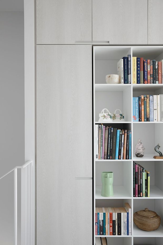 Close-up interior view of a built-in white and pale grey storage wall combining a tall concealed cabinet with open, square-cubed bookshelves. The shelves contain books and a few decorative objects, while a white stair balustrade is visible at the left edge. The joinery has a minimalist, clean-lined appearance with subtle light wood grain and slim horizontal handles.