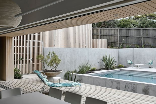 A refined contemporary outdoor entertaining area with a timber-lined covered terrace, pale timber decking, a raised rectangular swimming pool, and concrete retaining walls. Vertical timber screening, landscaped planting, and a privacy fence frame the pool, while outdoor dining furniture and a patterned lounge chair create a relaxed resort-like setting.