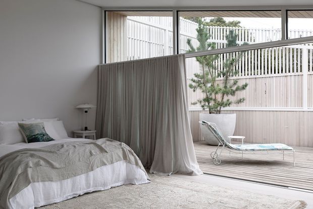 A bright, serene bedroom with a low-profile bed dressed in neutral linens, a soft draped fabric divider, and full-height glazing opening to a private timber-decked courtyard. The courtyard includes a large planter, lounge chair, and pale vertical privacy screening. The interior is restrained and contemporary with a soft neutral palette and abundant natural light.