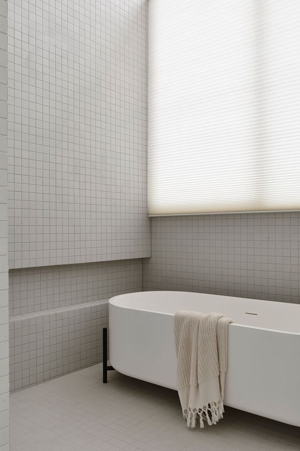 A minimalist bathroom featuring a freestanding white bathtub against walls covered in small square light-grey tiles. A white pleated window blind filters daylight above the tub, while a neutral woven towel hangs over the bath edge. The space has a clean, restrained, monochromatic appearance with subtle black tub supports.