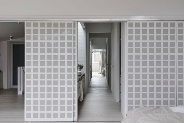 A bright, minimalist bedroom view looking through a central opening into a long hallway. Two large white grid-pattern sliding screens or room dividers frame the passage, with pale timber-look flooring, white walls, and a glimpse of a console and potted plant along the corridor. The edge of a bed is visible in the foreground, confirming the camera is positioned in a bedroom.