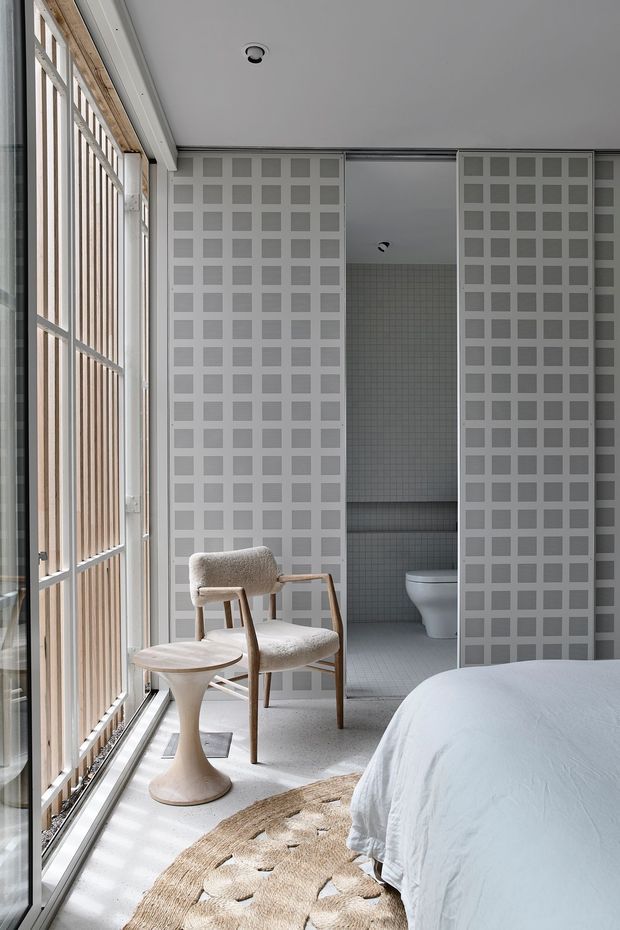 A bright, minimalist bedroom with a partially visible bed, upholstered mid-century-style armchair, sculptural side table, woven rug, and full-height glazed/slatted wall. A bathroom is directly connected to the bedroom through sliding patterned privacy screens, with a toilet and tiled walls visible. The room uses a restrained grey, white, and light timber palette with strong geometric screening.