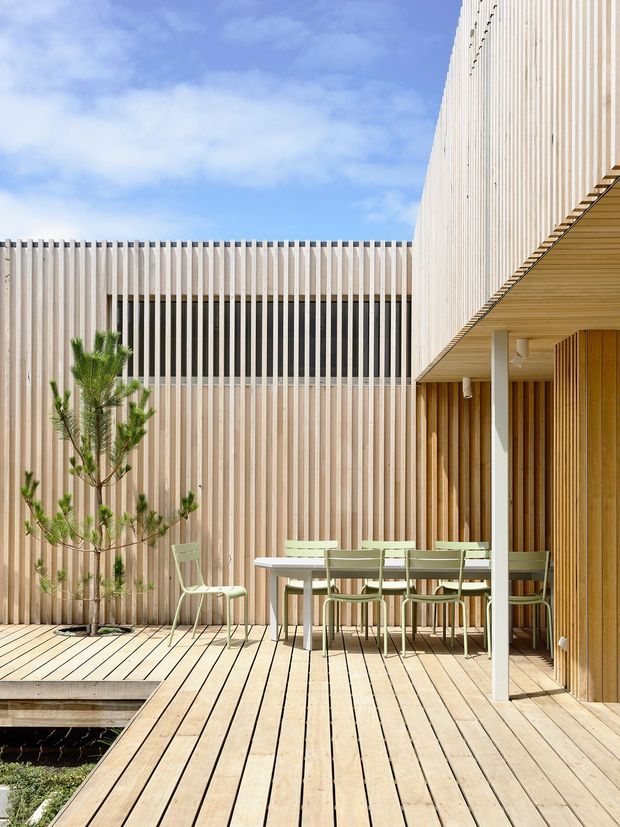 A bright contemporary courtyard terrace enclosed by vertical pale timber screening and cladding. A timber deck surrounds a small recessed water feature, with a light outdoor dining table and green chairs beneath a covered timber-lined overhang. The minimalist architectural composition uses strong vertical and horizontal lines, with a young sculptural tree adding greenery.