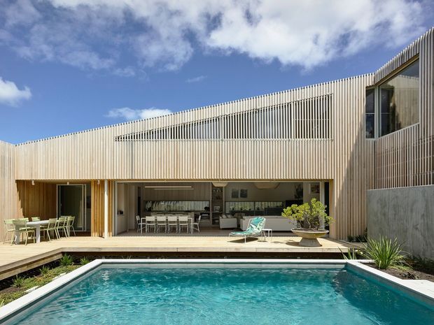 Contemporary two-storey residence clad in closely spaced light timber battens, opening onto a broad timber deck and rectangular swimming pool. The open-plan ground floor appears to contain a kitchen, dining and living area, while the upper level features screened glazing and a projecting terrace or balcony. Bright blue sky, restrained landscaping and clear pool water create a refined indoor-outdoor setting.