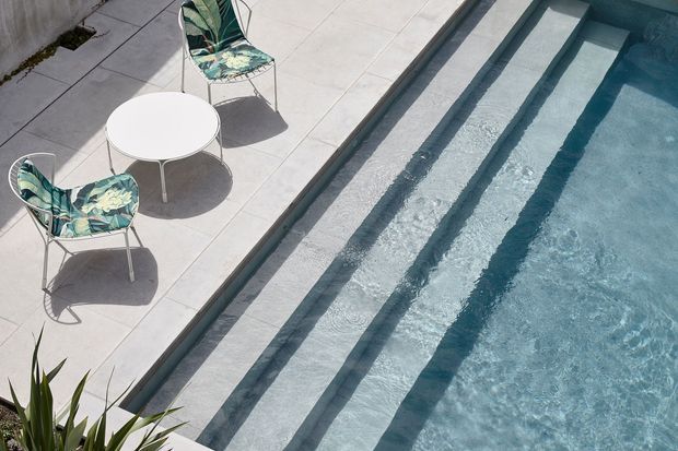 A sunlit pool area viewed from above, featuring pale tiled paving, broad submerged entry steps, and clear blue water. Two patterned outdoor lounge chairs and a small round white table sit beside the pool, with a small amount of planting visible at the edge.