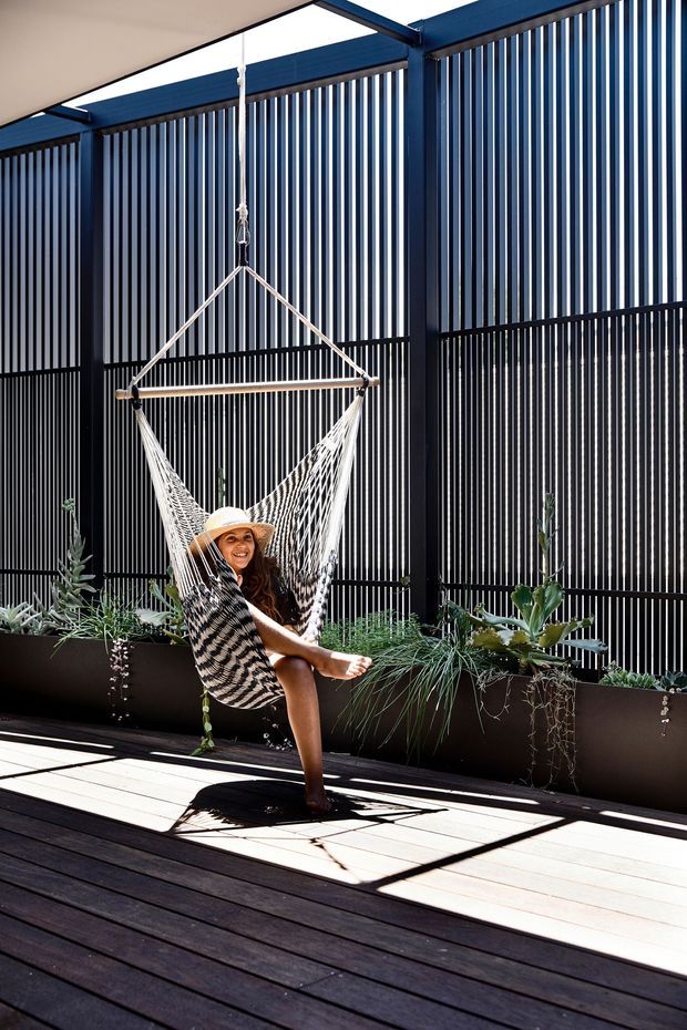 A bright outdoor terrace or balcony with dark timber decking, deep blue-black metal framing, and vertical slatted screening. Long built-in planter boxes filled with succulents and grasses line the perimeter, while a hanging woven hammock chair occupies the centre. Strong sunlight creates pronounced linear shadows across the deck, giving the space a relaxed contemporary indoor-outdoor character.