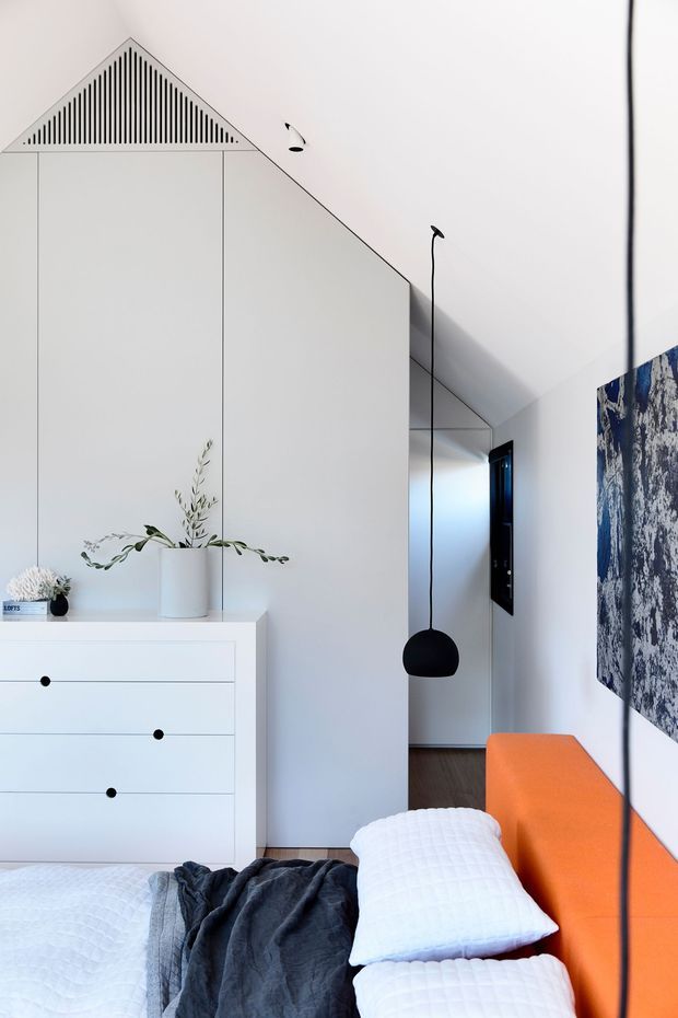 Bright minimalist bedroom with a steeply sloped white ceiling and integrated triangular vent detail. The room features a white built-in dresser, a bed with white quilted bedding and dark throw, a vivid orange upholstered bedhead or bench-like feature, a black pendant light, dark timber flooring, and a large abstract monochrome artwork.