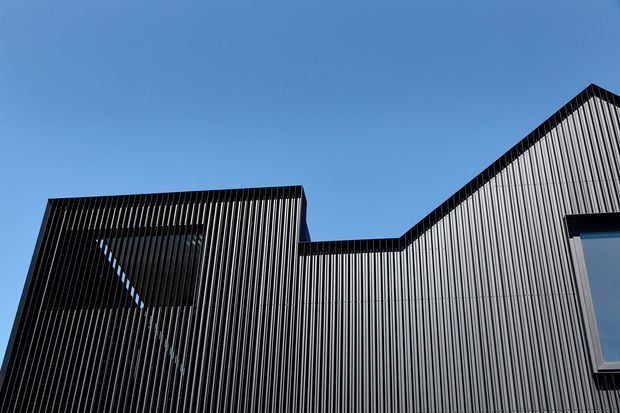 Close-up upward view of a modern building facade clad in closely spaced vertical dark grey metal fins or battens. The angular roofline includes a stepped low section and a steep gabled form, with tall reflective glazing partially visible behind the screen. A clear blue sky provides the background.