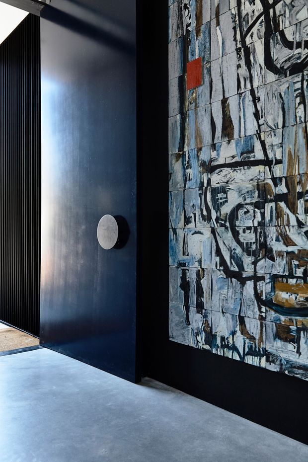 A dramatic, minimalist interior detail featuring a deep black-blue metal or painted wall panel with a circular pull or handle, a narrow black vertical screen or slatted opening, and a large abstract artwork mounted on a dark wall. The floor is smooth grey concrete, creating a restrained industrial gallery-like atmosphere with strong black, grey, white, and blue tones.