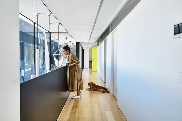 A bright, minimalist corridor in a contemporary interior, featuring pale timber flooring, white walls and ceiling, full-height glass partitions with dark metal framing, and a long dark built-in counter or balustrade. A vivid yellow door provides a small accent of colour. The space appears to be a modern workplace or studio, with a person and dog present.