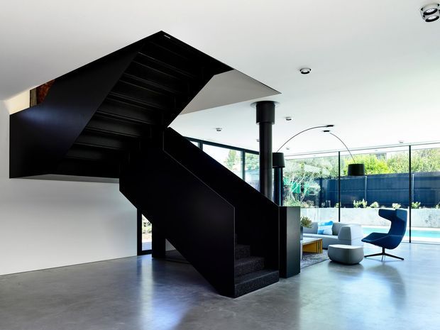 A dramatic modern open-plan interior dominated by a sculptural black steel staircase with angular folded geometry. The space has white walls and ceiling, polished grey concrete flooring, black structural columns, extensive floor-to-ceiling glazing, and a minimalist seating area overlooking a pool and landscaped courtyard. Recessed downlights and a curved floor lamp contribute to the clean contemporary atmosphere.