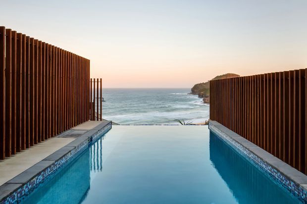 A narrow rectangular infinity pool leads toward a coastal ocean view at sunset. Tall vertical timber privacy screens line both sides, with pale stone paving and blue patterned pool-edge tiling. A rocky headland and waves are visible beyond the pool, creating a secluded, modern seaside setting.
