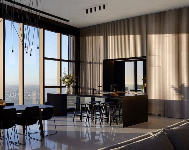 A sophisticated open-plan kitchen and dining area with floor-to-ceiling windows overlooking a cityscape. The space features a dark island with bar seating, vertically slatted timber wall panelling, glossy pale flooring, black-framed glazing, pendant lighting, and a large reflective opening above the kitchen bench. A dining table and sofa are visible in the foreground.