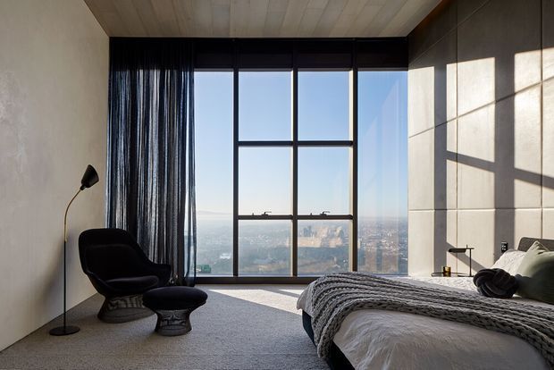 A serene contemporary bedroom with floor-to-ceiling black-framed glazing overlooking a distant cityscape. The room features a low bed with layered grey and white bedding, a black sculptural lounge chair and ottoman, dark sheer curtains, a pale carpet, a timber-lined ceiling, and large-format light stone or concrete wall panels illuminated by strong sunlight.