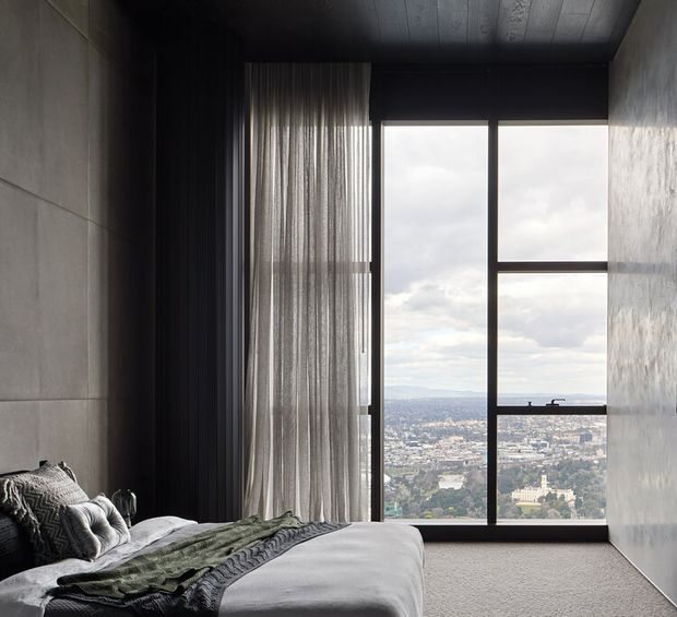Moody contemporary bedroom with a low-profile bed in the foreground, layered grey and green bedding, floor-to-ceiling sheer curtains, dark window framing, textured wall surfaces, and a large glazed opening overlooking a cityscape and distant hills. The room has a restrained, refined palette and strong vertical proportions.