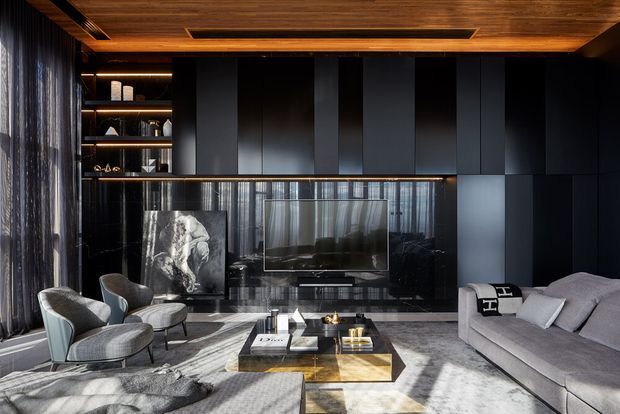 A sophisticated dark-toned living room with a large sectional sofa, sculptural lounge chairs, a central coffee table, built-in shelving, a wall-mounted television, and floor-to-ceiling curtains. The room features black wall panelling, reflective dark surfaces, marble-look finishes, and a richly timber-lined ceiling with recessed linear lighting.