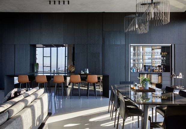 A sophisticated open-plan living, dining and kitchen space with dark vertically ribbed wall panelling, a black kitchen island with warm wood bar stools, a glass-fronted drinks cabinet, contemporary dining furniture, and sculptural pendant lights. Strong daylight enters through large windows and reflects across the polished floor, creating a refined, dramatic atmosphere.