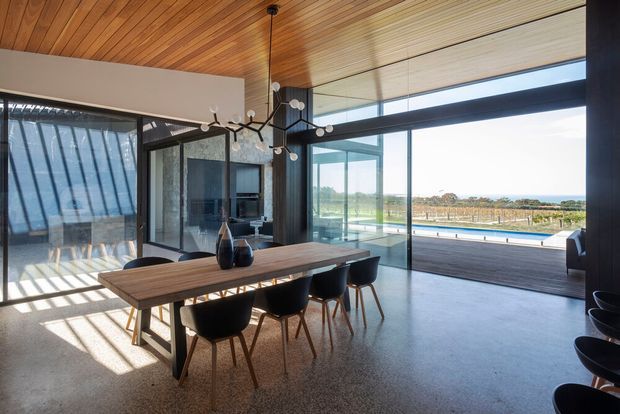 Contemporary open-plan dining area with a long timber dining table and dark upholstered chairs beneath a sculptural pendant light. The room features extensive floor-to-ceiling glazing, a warm timber-lined ceiling, dark-framed glass walls, polished concrete flooring, and views toward a pool and open landscape.